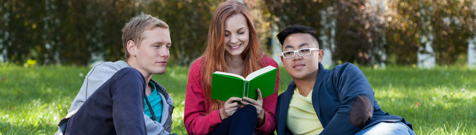 Two male students and one female student sitting on the grass reading a green book